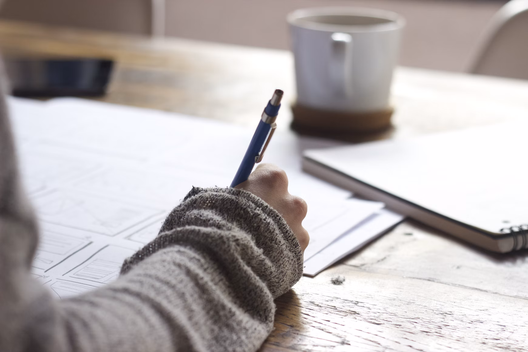 Person writing on paperwork at a wooden table with a notebook, coffee mug, and laptop nearby