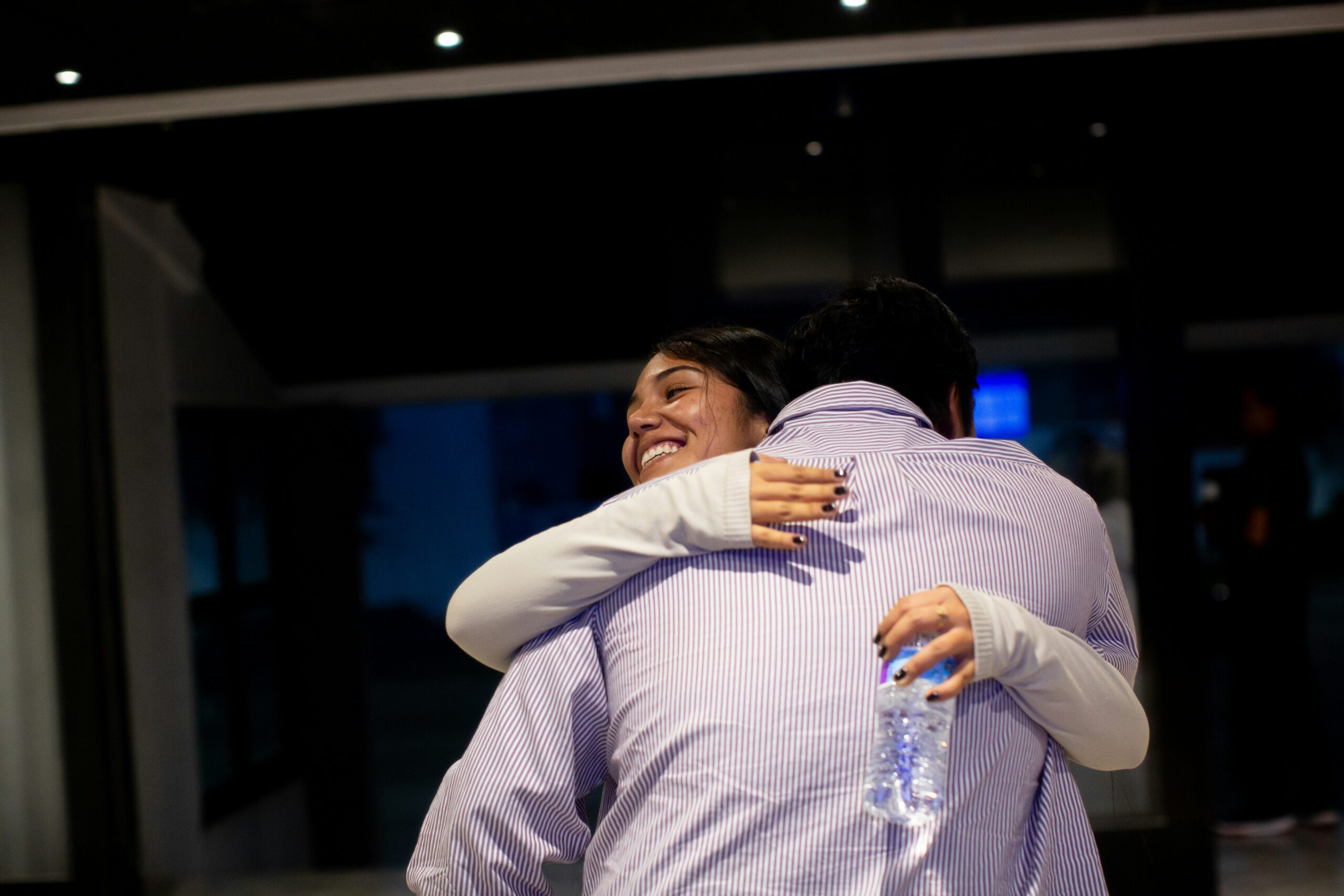A man and a woman hug. She is facing us, smiling, and holding a water bottle. He is facing away from us and ss wearing a striped Oxford-style shirt.